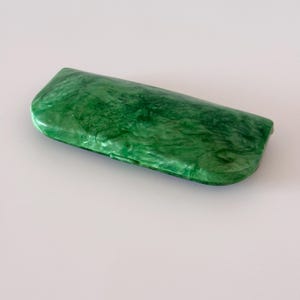Pode incluir: Um estojo rígido retangular para óculos com um padrão de mármore verde esmeralda vibrante, apresentando tons claros e escuros em espiral. O estojo possui um acabamento liso e brilhante com uma forma curva e arredondada para proteger os óculos.