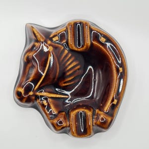 May include: A brown ceramic ashtray shaped like a horse head. The ashtray has a glossy finish and features a horseshoe design. The horse's mane and facial features are detailed with lighter brown accents. The ashtray is a vintage item.