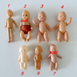May include: A selection of seven vintage baby dolls in different poses. The dolls are made of plastic and have a light skin tone. Some dolls are wearing red shorts. Two dolls have the word "sold" written on them.