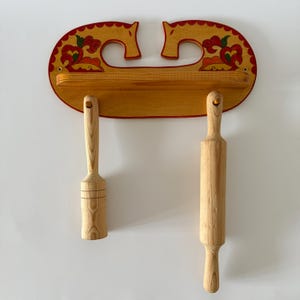 Puede incluir: Juego de utensilios de cocina de madera con un rodillo y un mortero colgando de un estante decorativo. El estante está pintado de amarillo con detalles rojos y florales. El rodillo y el mortero son de madera clara.