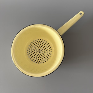 May include: A yellow enamel strainer with a long handle. The strainer has a round bowl with small holes for draining liquids. The rim and handle are outlined in black. The handle has a hole for hanging.