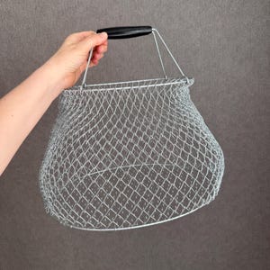 Puede incluir: Una cesta de malla de alambre plateada con un asa negra. La cesta tiene un fondo redondeado y un asa que se arquea hacia arriba. La malla está hecha de pequeñas aberturas en forma de diamante.