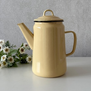 Könnte beinhalten: Eine cremefarbene Emaille-Kaffeekanne mit schwarzem Rand und Griff. Die Kanne hat einen Ausguss, einen Deckel und einen Griff. Sie steht auf einer weißen Oberfläche. Ein klassischer Küchenartikel.
