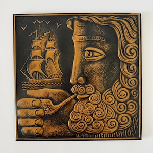 May include: A square wall plaque featuring a relief design in copper and black tones. The artwork depicts a profile of a bearded figure smoking a pipe, with a sailing ship on the water in the background. The surface has a textured, metallic finish.