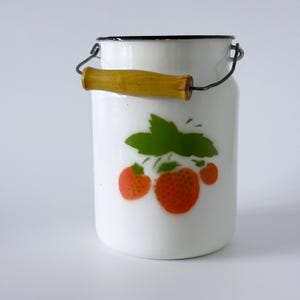 Könnte beinhalten: Weißer Emaille-Milchkrug mit schwarzem Rand und Holzgriff. Der Krug zeigt ein Erdbeermuster mit grünen Blättern und roten Beeren. Der Griff hat eine hellbraune Farbe.