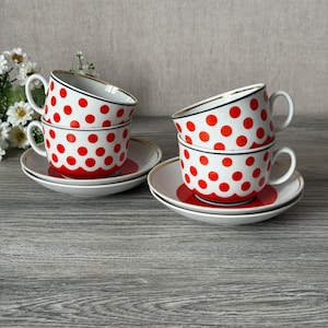 May include: Four stacked teacups and saucers. The white cups feature red polka dots, black trim, and gold rims. The saucers have a red center and white border. The cups are on a gray wooden surface.