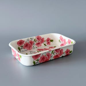 Może przedstawiać: Prostokątna biała emaliowana taca z kwiatowym wzorem. Taca ma różowe kwiaty z zielonymi liśćmi i szarymi akcentami. Kwiaty są rozmieszczone na powierzchni tacy.