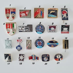 May include: A collection of twenty-one vintage Soviet space-themed metal enamel pins arranged in four rows. The pins feature various shapes, including shields, rectangles, circles, and ovals, displaying imagery of cosmonauts, rockets, satellites, and lunar rovers with Cyrillic text and dates.