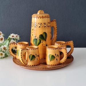 May include: A wooden pitcher with a lid and four matching mugs, all with carved handles and birch leaf designs. The set is displayed on a round wooden tray. The pitcher and mugs are light brown with green and black accents.