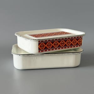 May include: Two rectangular white enamel storage containers stacked on top of each other. The top container features a decorative band with a geometric pattern in red, orange, black, and yellow. Both containers have a thin black rim along the edges.