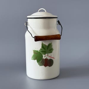 Pode incluir: Jarro de leite esmaltado creme com aro e alça pretos. O jarro apresenta uma alça de madeira e um desenho pintado de folhas verdes e bagas vermelhas. Um item de decoração de cozinha vintage.