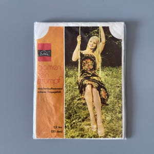May include: A packet of Erda brand pantyhose. The packaging features a woman on a swing wearing a floral dress. The package has German text, including "Damen Fein Strumpf" and "2.2 tex (20 den)." The packaging is orange and white.