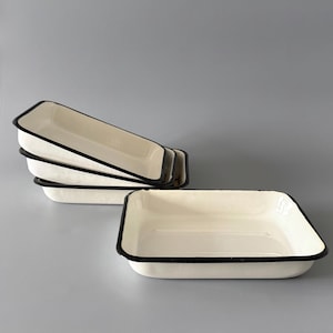 Puede incluir: Un conjunto de bandejas rectangulares de esmalte blanco roto con ribetes negros. Una bandeja está en primer plano. Las bandejas están vacías y parecen nuevas. El fondo es de color gris liso.