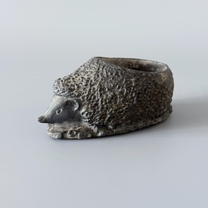 May include: A small, silver-coloured hedgehog-shaped container. The container has a textured body and a smooth, pointed snout. The opening is a small, round cavity. The hedgehog is a decorative item.