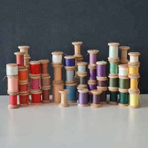 May include: A collection of wooden spools of thread in various colors, including red, blue, green, yellow, and white. The spools are arranged in a row on a dark surface.
