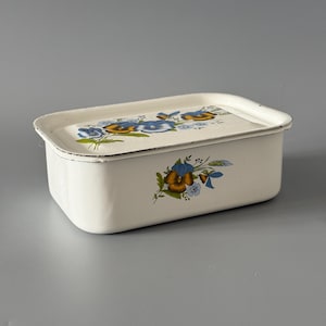Peut inclure: Une boîte de rangement rectangulaire en émail blanc avec un couvercle assorti. L'extérieur et le couvercle présentent un motif floral de pensées bleues et brunes avec des feuilles vertes. Les bords sont soulignés d'un fin liseré noir.