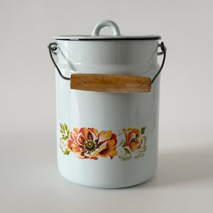 Puede incluir: Un recipiente de esmalte azul claro con tapa y un asa metálica con empuñadura de madera. El contenedor cilíndrico está decorado con una banda horizontal de ilustraciones florales en naranja, amarillo y verde en la parte inferior.