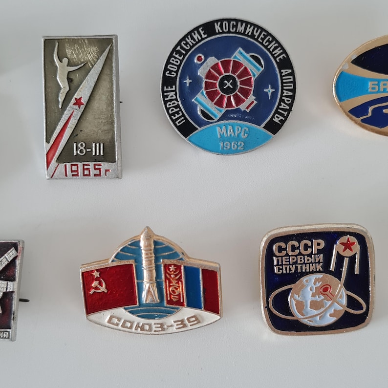 May include: A collection of six vintage Soviet space-themed enamel pins. The pins feature various designs including a rectangular 1965 badge, a circular Mars 1962 emblem, a Soyuz-39 mission pin with flags, and a square CCCP Sputnik commemorative badge.