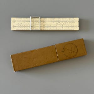 May include: An ivory-colored slide rule with a silver frame and a brown rectangular case. The slide rule has multiple scales and markings. The case has a circular marking on it.