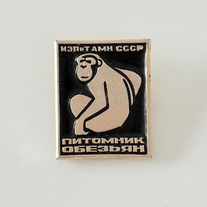 Puede incluir: Un pin de esmalte negro y plateado con un contorno blanco. El pin presenta una imagen de dibujos animados de un mono sentado. El texto "ИЗПИТ АМН СССР" está en la parte superior del pin y el texto "ПИТОМНИК ОБЕЗЬЯН" está en la parte inferior del pin.