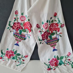 May include: White fabric with embroidered floral designs. The embroidery features red and pink roses, green leaves, and colorful vases. The fabric is folded, displaying the detailed floral patterns.