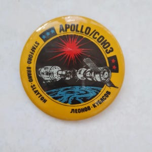 May include: A yellow round pin with a black and white image of two spacecraft docked together in space. The text "APOLLO/СОЮЗ" is written in a circle around the image. The text "STAFFORD - BRAND - SLAYTON" is written on the left side of the pin and the text "ЛЕОНОВ - КУБАСОВ" is written on the right side of the pin.