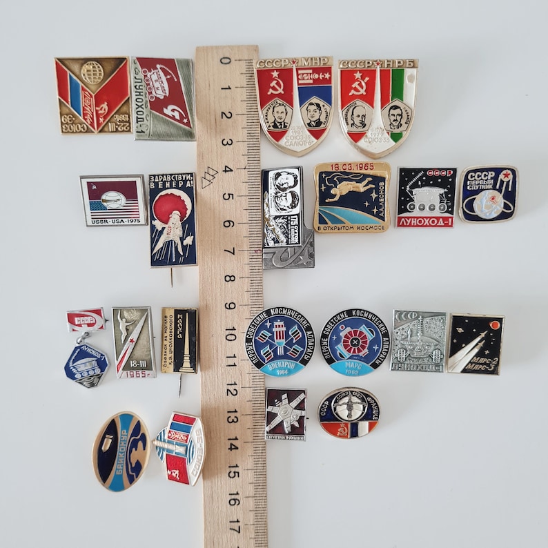 May include: A collection of twenty-four vintage Soviet space-themed metal enamel pins arranged around a wooden ruler measuring 17 centimeters. The pins feature various shapes, including shields, circles, and rectangles, displaying imagery of rockets, cosmonauts, planets, and Cyrillic text.