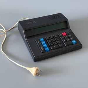 Könnte beinhalten: Ein schwarzer Vintage-Taschenrechner mit grünem Display und einer Tastatur mit blauen, roten und schwarzen Tasten. Er ist mit einem cremefarbenen Netzkabel und Stecker verbunden. Das Gerät befindet sich auf einer grauen Oberfläche.