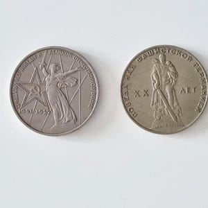 May include: Two silver commemorative coins. One coin features a woman holding a sword with text in Cyrillic script and the dates 1941-1945. The other coin depicts a soldier carrying a child, also with Cyrillic text.