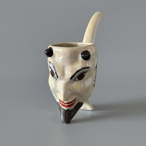 May include: A ceramic devil-faced creamer with a pearlescent finish. The creamer features black horns, red lips, and detailed facial features. A curved handle extends from the back, and the creamer stands on a small foot.