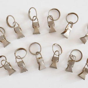 May include: A set of 12 silver metal curtain clips with rings. Each clip has a spring-loaded mechanism to hold curtains in place.