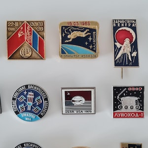 May include: A collection of eight vintage Soviet space-themed enamel pins featuring various designs, including the Soyuz 39 mission, A.A. Leonov in space, Venus exploration, the Electron satellite, the 1975 USSR-USA mission, and the Lunokhod-1 lunar rover.
