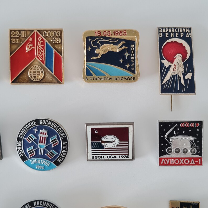 May include: A collection of eight vintage Soviet space-themed enamel pins featuring various designs, including the Soyuz 39 mission, A.A. Leonov in space, Venus exploration, the Electron satellite, the 1975 USSR-USA mission, and the Lunokhod-1 lunar rover.