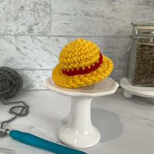 May include: A small yellow crocheted hat with a red band. The hat is sitting on a white cake stand.