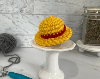 May include: A small yellow crocheted hat with a red band. The hat is sitting on a white cake stand.