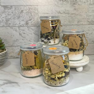 May include: Four glass jars filled with herbs, spices, and salts, each labeled with a different word: Protection, Prosperity, Creativity, and Attract Romance. The jars are displayed on a white surface with a succulent plant in the background.