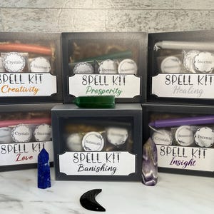 May include: Six spell kits in black boxes, each labeled with a different intention: Creativity, Prosperity, Healing, Love, Banishing, and Insight. Each kit contains a candle, crystals, incense, and botanicals. Various crystals and a black crescent moon are also present.