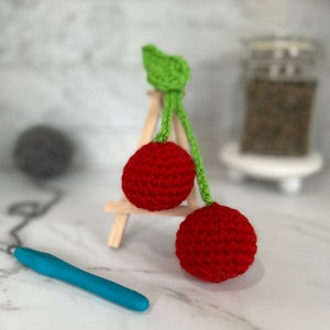 May include: Two red crocheted cherries with green crocheted stems and leaves. The cherries are on a small wooden easel.