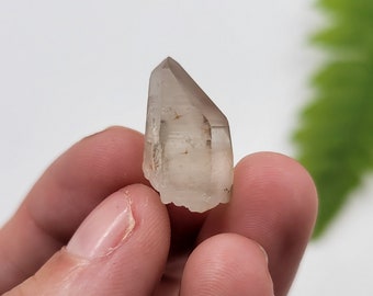May include: A clear quartz crystal point with a slight brown tint. The crystal is pointed at the top and has a flat base. It is being held in a hand.