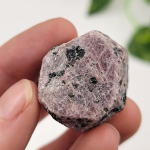 Könnte beinhalten: Ein roher, rosafarbener Rubin-Kristall mit schwarzen Einschlüssen. Der Kristall ist unregelmäßig geformt und hat eine natürliche, unpolierte Oberfläche.