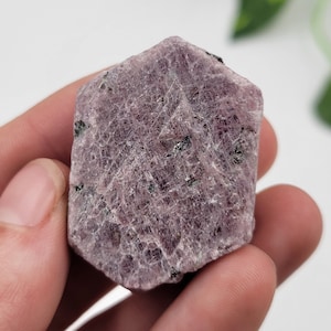 May include: A raw pink ruby crystal with black inclusions. The crystal is faceted and has a hexagonal shape.