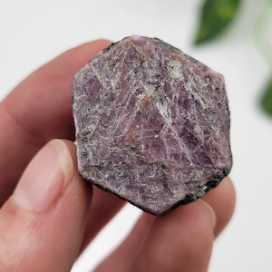 Puede incluir: Un cristal de rubí facetado de color rosa oscuro con una textura natural rugosa. El cristal tiene una forma hexagonal y un color profundo y rico.