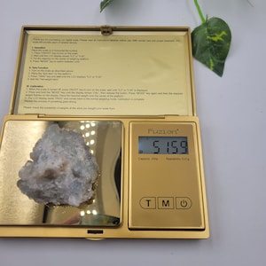 May include: A gold digital scale with a white crystal on the platform. The scale reads 5.159 grams. The scale has a capacity of 200 grams and a readability of 0.01 grams.