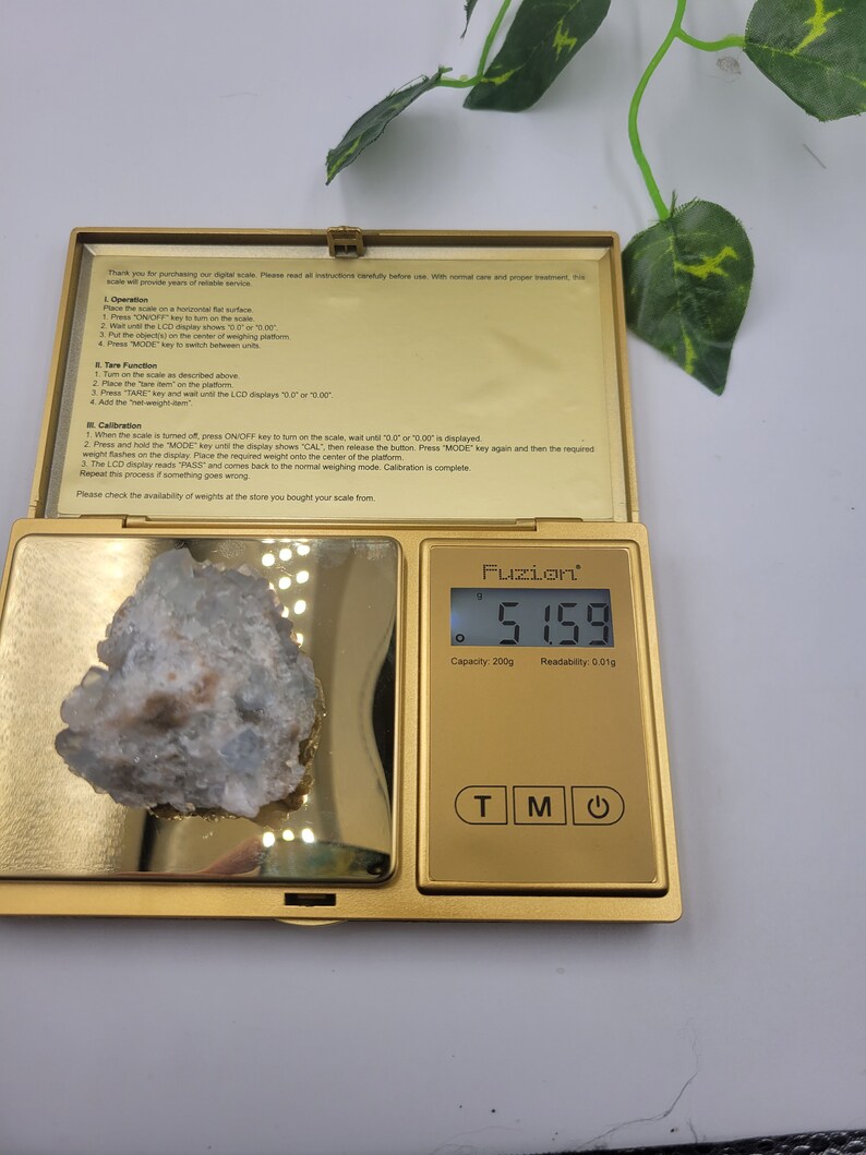 May include: A gold digital scale with a white crystal on the platform. The scale reads 5.159 grams. The scale has a capacity of 200 grams and a readability of 0.01 grams.