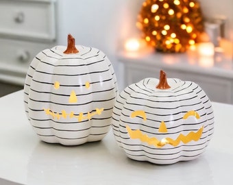 May include: Two white ceramic pumpkins with black stripes and carved jack-o'-lantern faces. The pumpkins have a warm glow coming from inside.