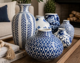 May include: A collection of blue and white ceramic vases with various patterns including leaf motifs, geometric diamonds, and floral designs. The vases are arranged on a wooden tray alongside two stacked books and a wooden bead garland.