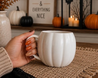May include: A white ceramic mug shaped like a pumpkin with vertical ridges and a curved handle. The mug has a smooth, glossy finish and a rounded body. It is held by a hand with multi-colored autumn-themed nail polish in shades of brown and orange.