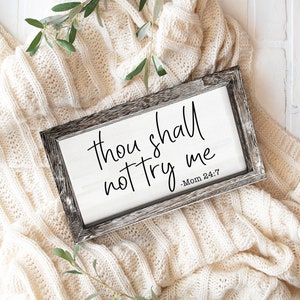 以下が含まれることがあります： 白い背景に「Thou shall not try me - Mom 24:7」という文字が書かれた素朴な木製の看板。看板は白いニットの毛布の上に置かれています。
