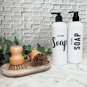 May include: Two white soap pump bottles with black pumps and black text that reads "Soap" and "Gia Roma" on a white countertop.  Two wooden scrub brushes are in a wooden tray.