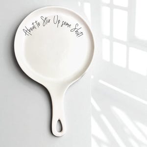 Op de afbeelding: Witte keramische lepelhouder met een handvat en de tekst "About to Stir Up some Shit" in zwart schrift. De lepelhouder is rond met een handvat en een gat aan het uiteinde.
